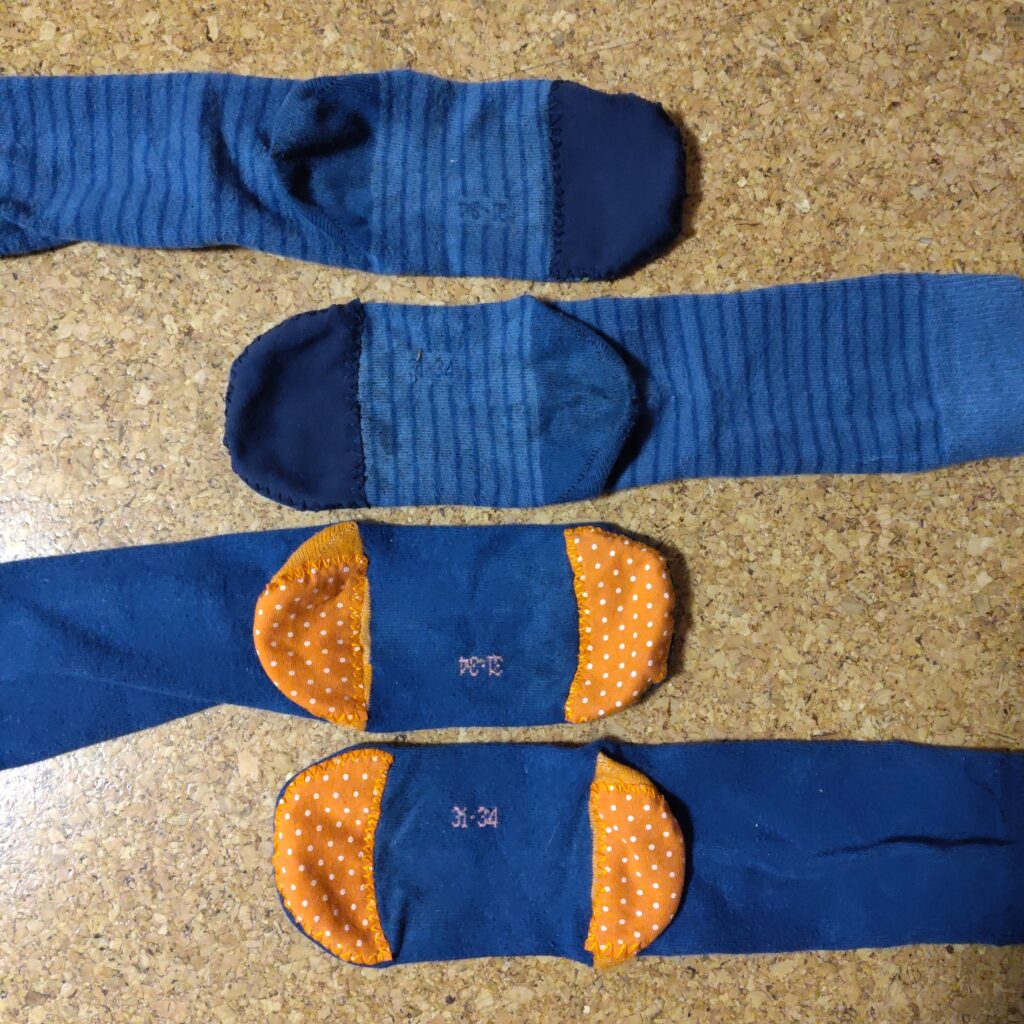 Aufnahme von zwei paar blauen Socken. Bei einem wurde der Zehenbereich mit blauem Stoff verstärkt. Bei dem zweiten Paar wurde Zehen- und Fersenbereich jeweils mit orangefarbenem Stoff verstärkt.